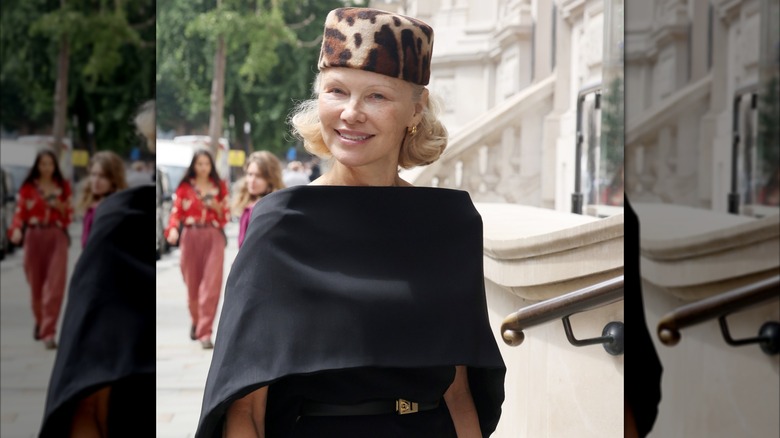Pamela Anderson smiling outside wearing a black caplet dress with a leopard-print pillbox hat.