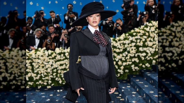 Rihanna posing at the 2025 Met Gala wearing a menswear-inspired outfit with a a floppy bow-tie and a large black hat.