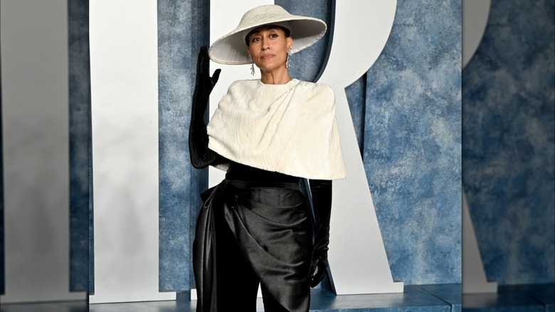 Tracee Ellis Ross posing at a red carpet event wearing a black outfit and gloves with a white caplet and a white hat.