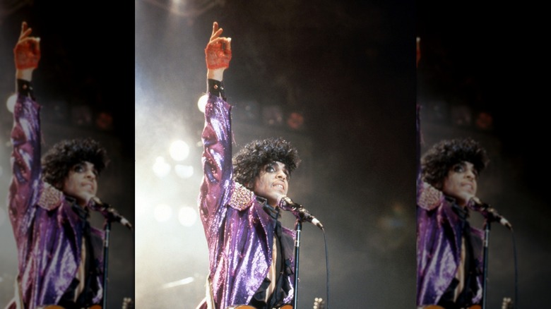 Prince performing during the Purple Rain tour wearing a sparkly purple jacket with his hand in the air.
