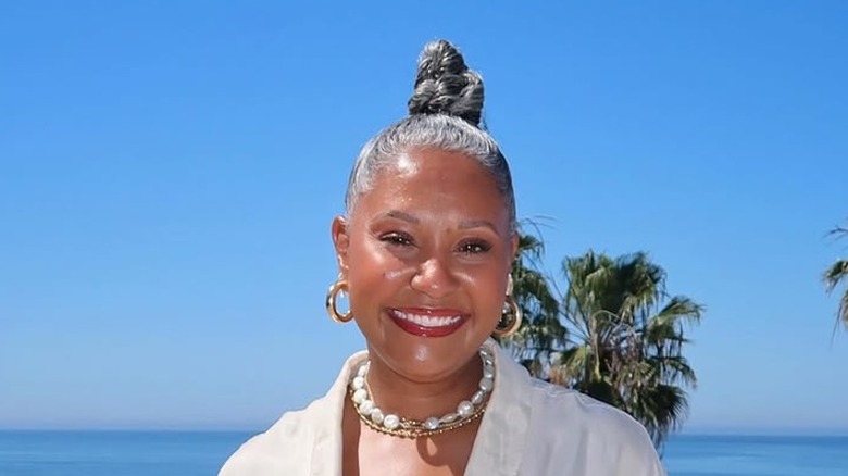 A woman with gray hair in a braided topknot wearing a white shirt, hoop earrings and a statement necklace.