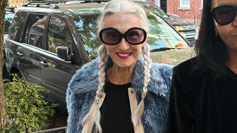 A woman with two long braids smiling on the street wearing a furry jacket and large sunglasses.