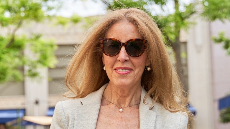 A woman with long hair wearing a tan blazer, pearl jewelry, and sunglasses.