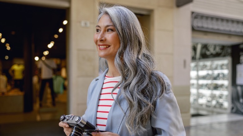 A woman with long, wavy hair smiling and holding a camera, wearing a red and white striped shirt and gray blazer.
