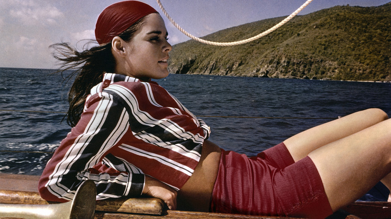 Ali MacGraw reclining on a sailboat wearing a red bandana, striped shirt, and red shorts.