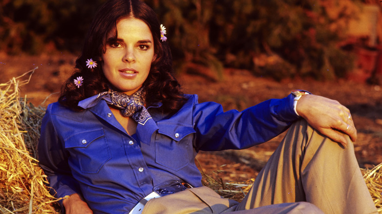 Ali MacGraw reclines on a bale of hay circa 1960s wearing a blue western-style shirt and bandana.