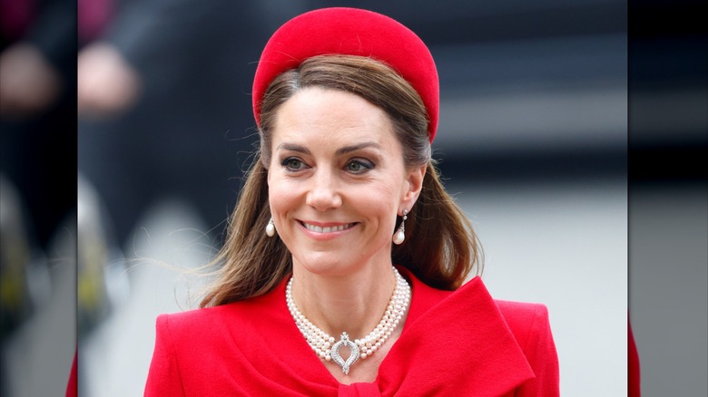 Kate Middleton smiles while walking on the street in a red outfit and a pearl choker around her neck