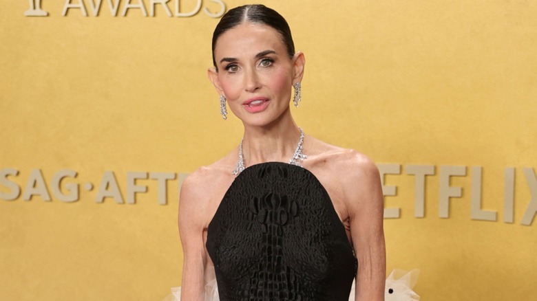 Demi Moore posing in a black halter dress with her hair slicked back and dangly earrings in front of a yellow background.