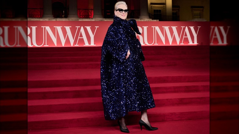 Meryl Streep on the red carpet wearing an oversized, dark-blue sparkling coat with simple black pumps and sunglasses.