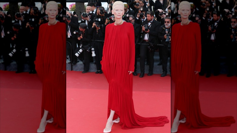 Tilda Swinton posing on the red carpet with her foot pointed and looking to the side wearing a flowing red dress.