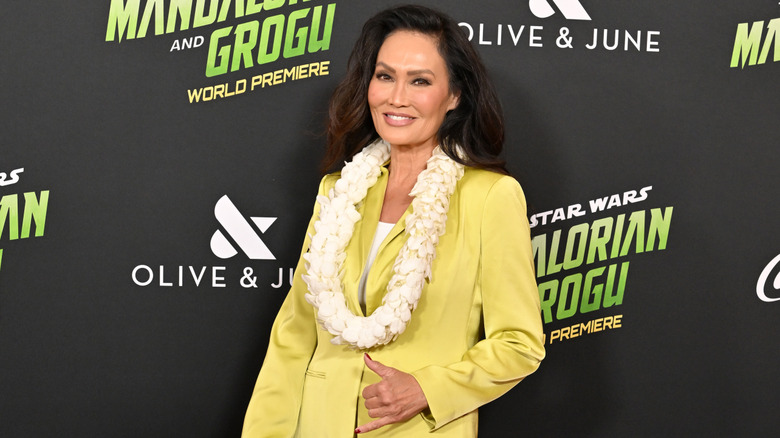 Tia Carrere throwing a shaka sign at a red carpet event wearing a light yellow suit and a lei.