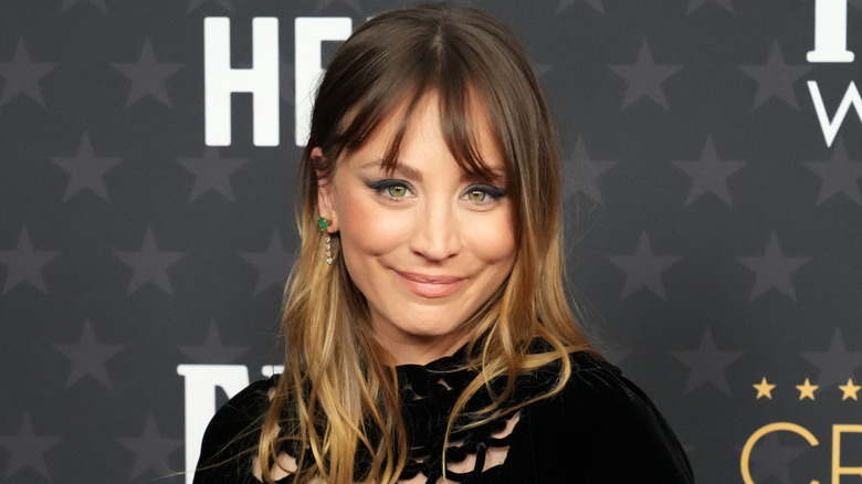 Kaley Cuoco smiling to the camera at a red carpet event while sporting dark eye makeup and a pale pink lip.