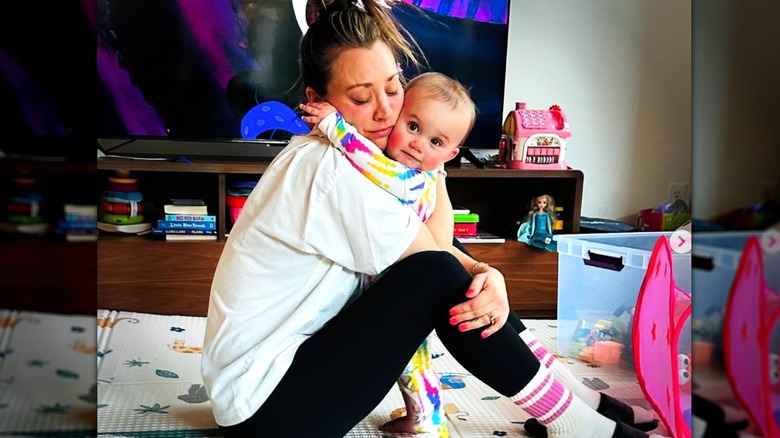 Kaley Cuoco wearing leggings and a T-shirt hugging her toddler daughter Matilda, sitting on a rug with toys around her.