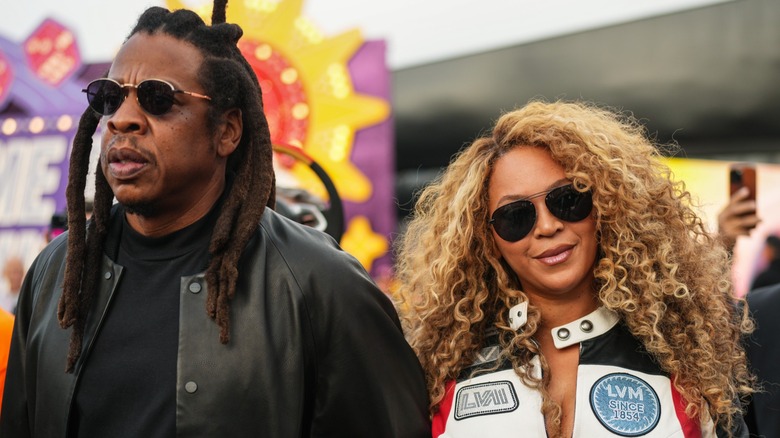 Jay-Z and Beyonce at an event. Jay-Z is a middle-aged man with brown skin and long locs. He is wearing black sunglassses and a black leather jacket. Beyonce is a woman with brown skin and long light brown curly hair. She is wearing dark aviator sunglasses and a black, white, and red motorcycle jacket.