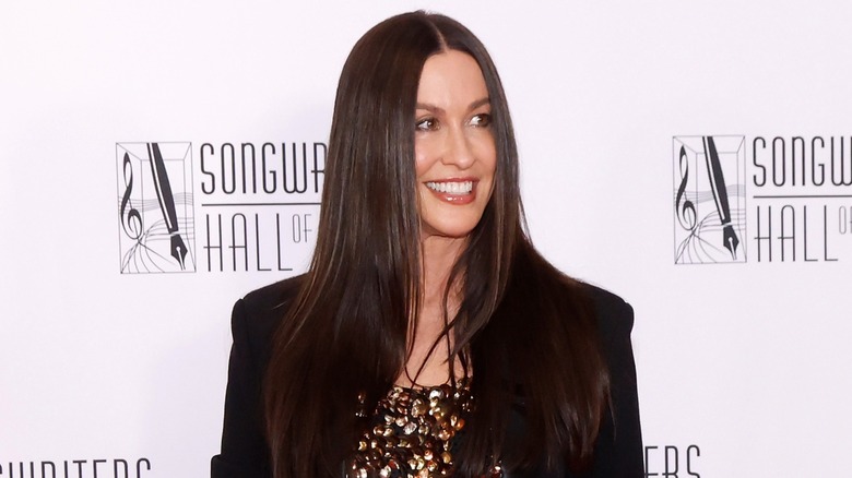 Alanis Morissette with straight hair wearing a sparkly gold top and black jacket on the red carpet.