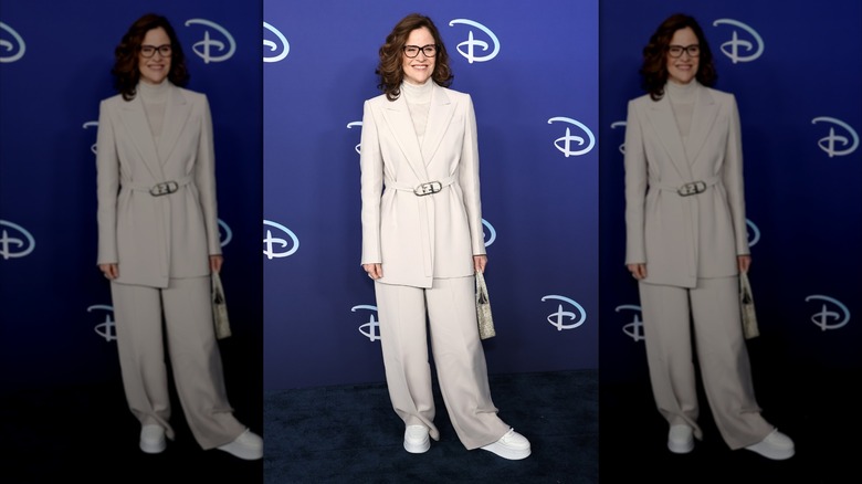 Ally Sheedy posing at an event in 2022 wearing an all-white suit and thick-framed glasses.