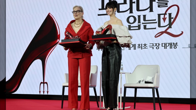 Meryl Streep and Anne Hathaway speak at a Korean "The Devil Wears Prada 2" promo event. Streep is an older woman with pale gray hair in a ponytail, black framed glasses, and a stylish red pantsuit. Hathaway is wearing her hair in a high bun, and an off-the-shoulder white top with bubble sleeves and black leather trousers.