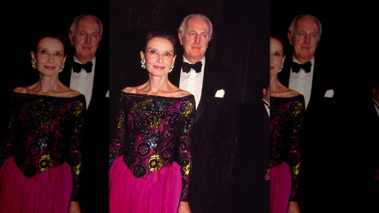 Audrey Hepburn in an elaborately-patterned off-the-shoulder dress and French fashion designer Hubert de Givenchy in a tuxedo at a fashion event.