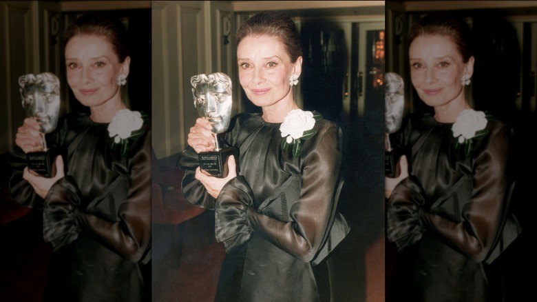 Audrey Hepburn posing with an award wearing a black gown with sheer sleeves and a white rose accent.