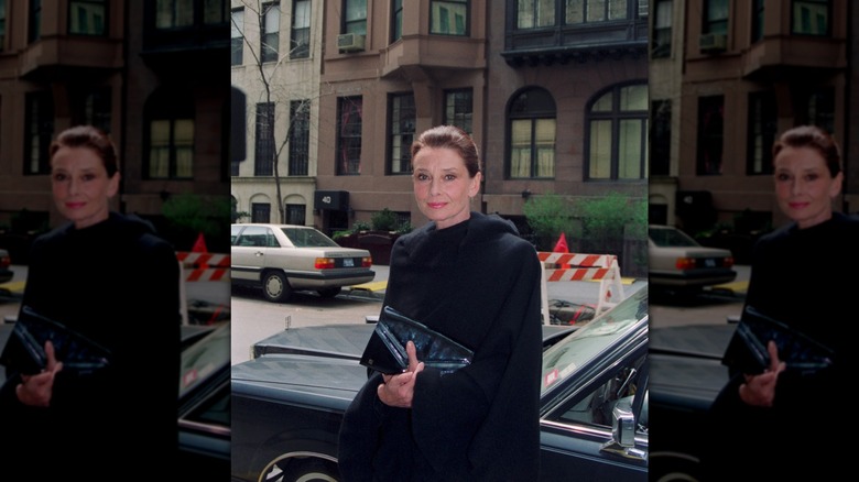 Audrey Hepburn on the street in New York City in the daytime wearing a black coat.