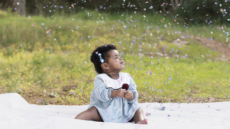 Baby girl sitting on a blanket, smiling with closed eyes as colorful confetti falls around her