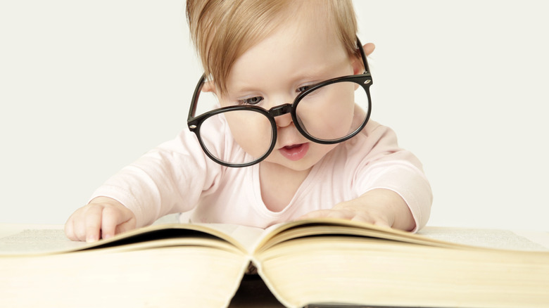 An infant wears oversized glasses while looking down at a thick book