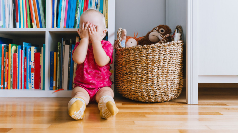 A toddler girl sits in front of a colorful bookshelf next to a basket of toys covering her face