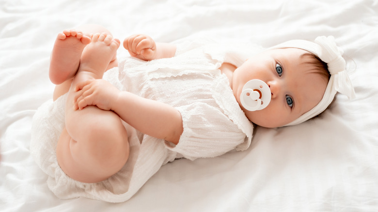 Baby girl with a pacifier dressed in white laying on a white blanket.