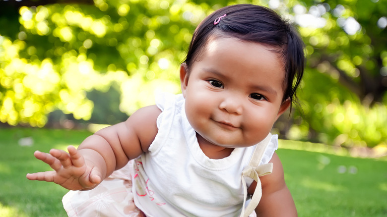 Smiling baby girl in a white outfit  and a pink barrette crawling on grass.