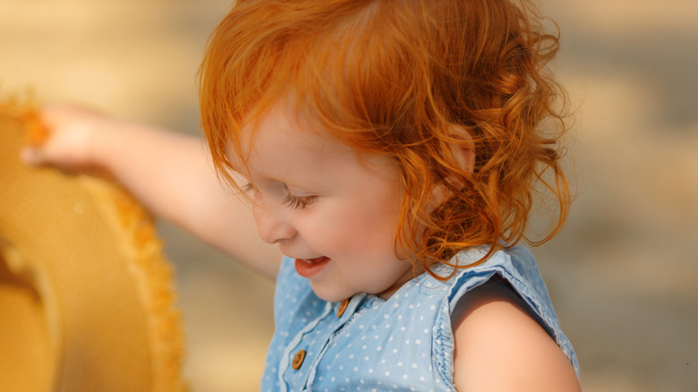 Smiling red-haired toddler wearing a blue-and-white polka dot top sitting outside.