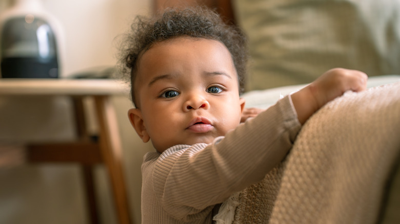 Baby boy wearing a ribbed shirt turned to the camera holding on to the side of a bed.