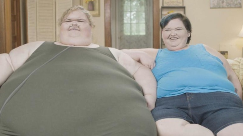 Sisters Tammy and Amy Slaton sit side by side on a couch. Tammy, pre-weight loss, wears a green dress and is using an oxygen tube while Amy is wearing a blue tank top and denim shorts