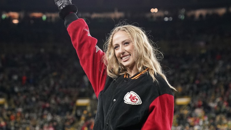 Brittany Mahomes waves to the crowd at the 2024 Super Bowl. Her blonde hair is down an wavy, with two small braids by her temples. She is wearing a Kansas City Chiefs-themed bomber jacket with a pair of black leather gloves.
