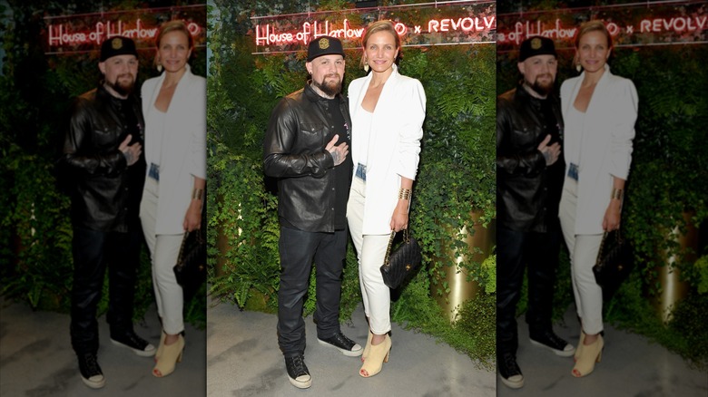 Benji Madden wearing an all-black outfit and hat posing in front of a leafy backdrop with Cameron Diaz wearing an all-white look.