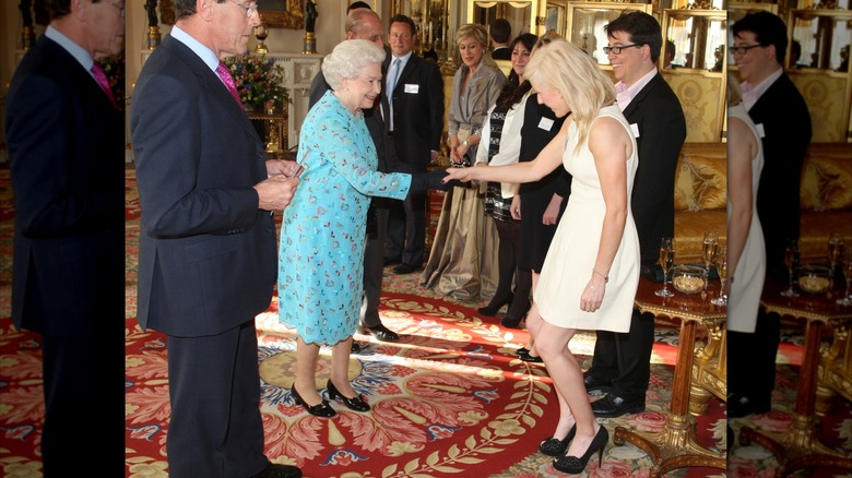Ellie Goulding wearing a white dress with black shoes curtsying and shaking hands with Queen Elizabeth wearing a blue dress.