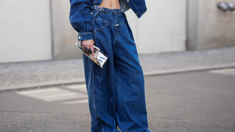 A woman wears low-rise baggy jeans and flip-flops while holding a Vogue magazine, her phone, and her sunglasses