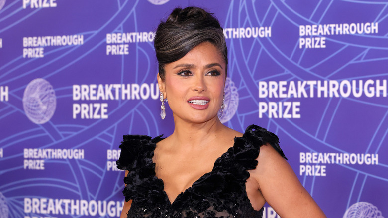 Salma Hayek posing at an event wearing a black gown with her hair in an updo with prominent gray streaks.
