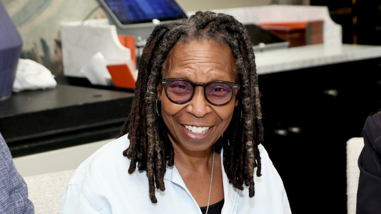 Whoopi Goldberg smiling at an event wearing a white shirt with black tinted glasses.