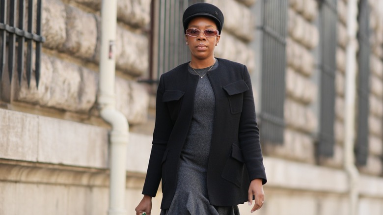 A woman walking in a dark-toned outfit, wearing a gray dress and black blazer