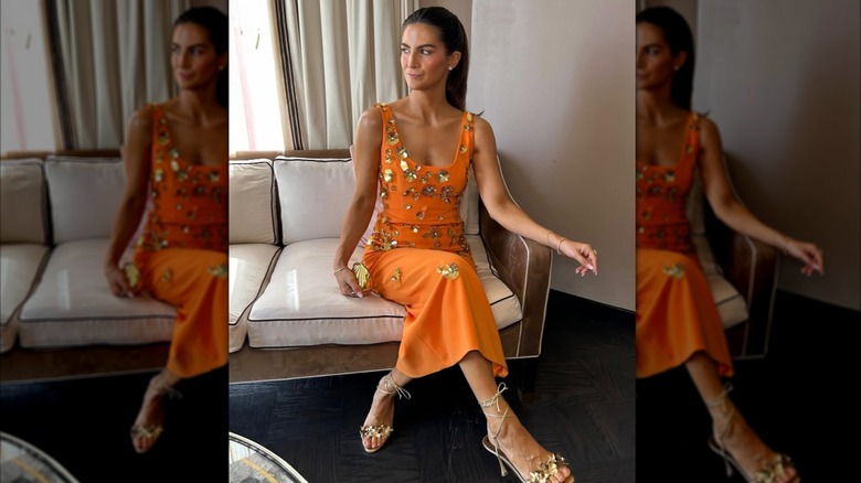 Kaitlan Collins sits cross-legged on a white and tan couch. She is wearing an orange dress covered in 3D gold floral appliques, gold strappy heels and minimal gold jewelry. Her hair is slicked back behind her shoulders
