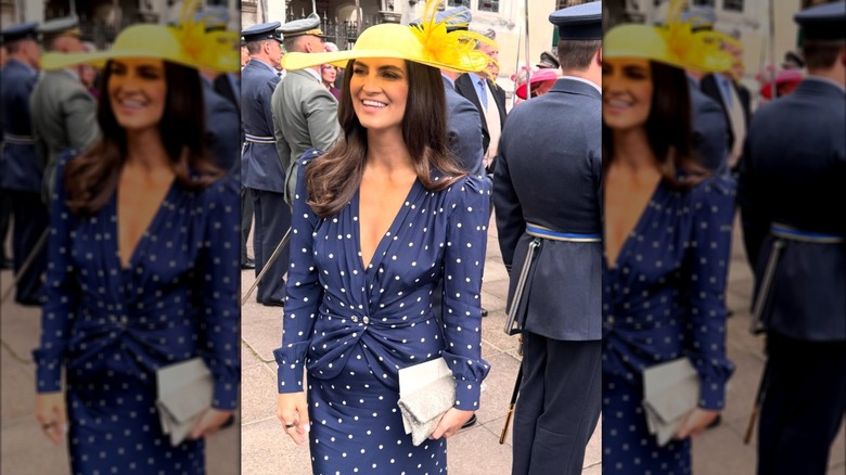 Collins is wearing a navy blue dress with white polka dots and a bright yellow wide-brimmed hat with a large yellow feather.