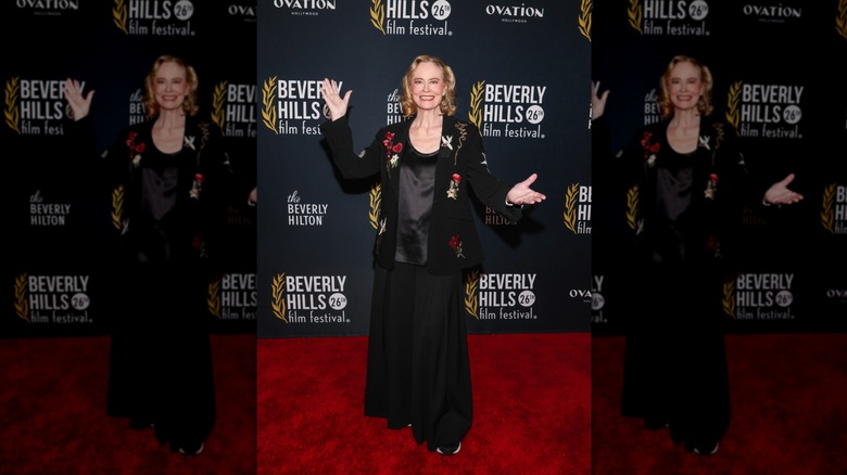 Cybill Shepherd smiles with open arms wearing a floral black blazer at a red carpet event.