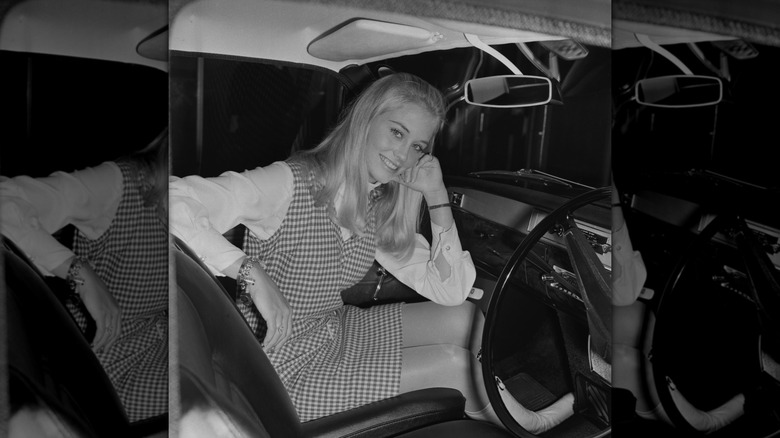 A black and white photo of Cybill Shepherd sitting in a car wearing a checked dress.