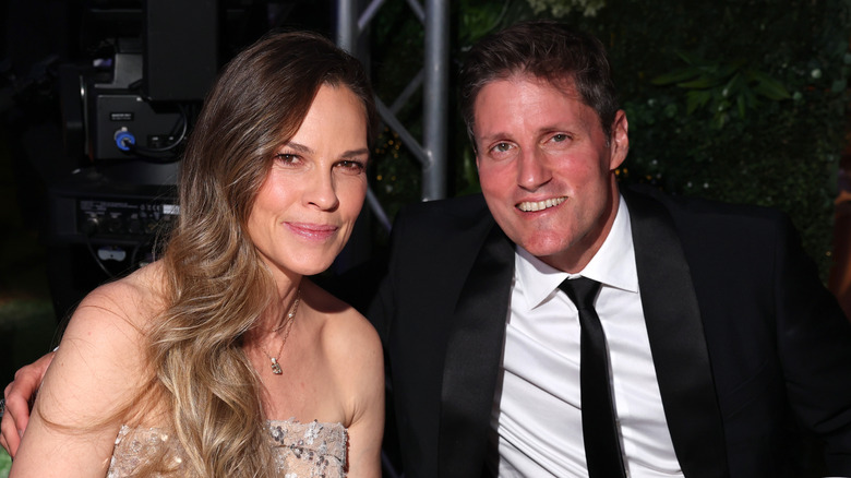 Hilary Swank smiling in a strapless dress next to husband Philip Schneider in a suit.