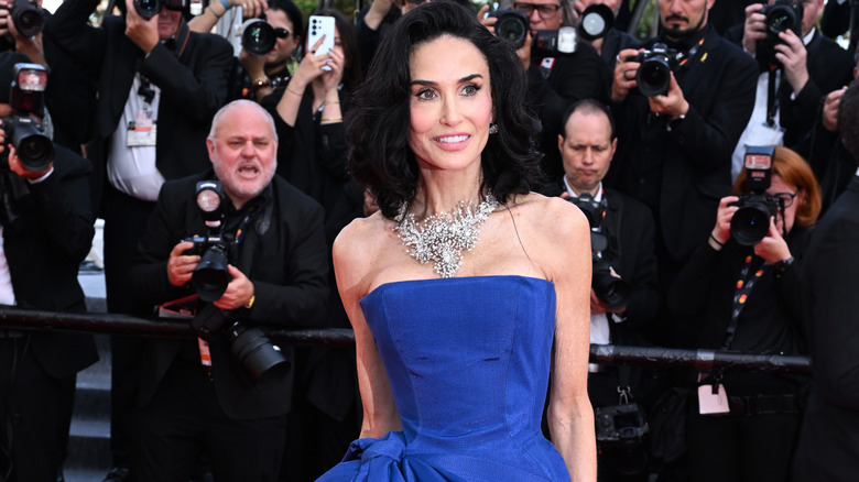 Demi Moore posing on the red carpet wearing a blue strapless gown and an ornate necklace in 2026.