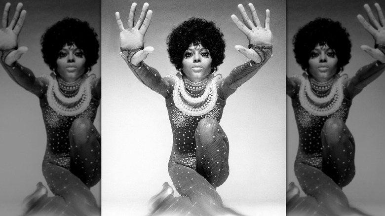 Diana Ross posing for a black and white portrait with her hands facing forward, wearing a black fishnet bodysuit covered in rhinestones.