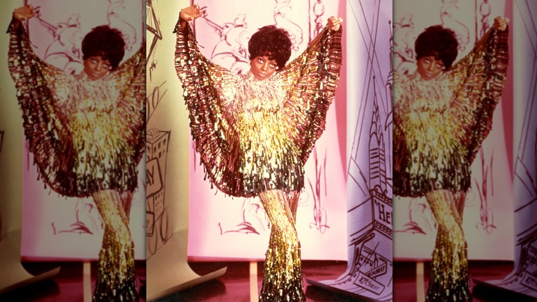 Diana Ross posing dramatically with her arms up wearing a sparkling gold outfit.