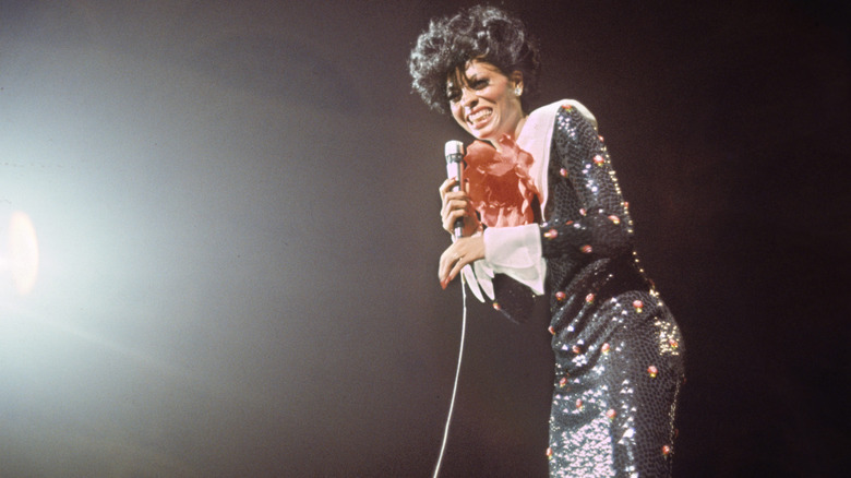 Diana Ross pictured on stage with a microphone wearing a black sequin dress with a broad white collar.