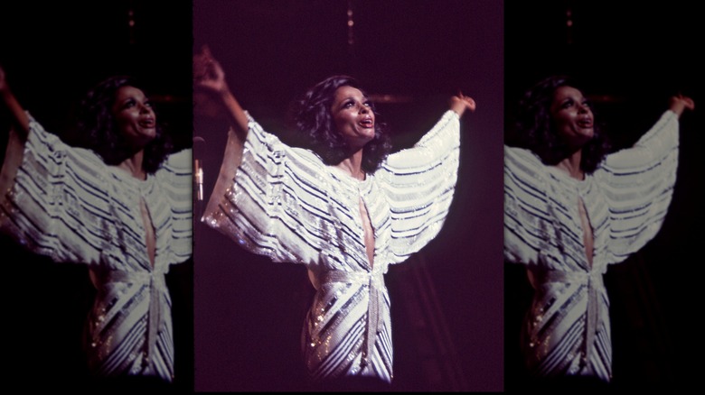 Diana Ross performing with her arms in the air wearing a white and black striped flowing gown.