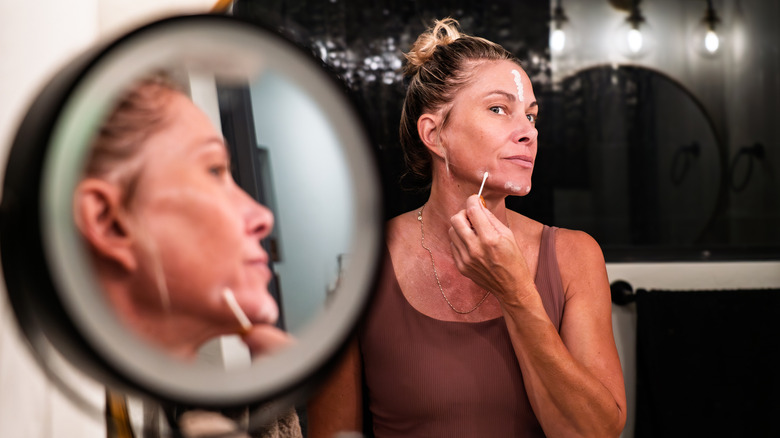 Middle-aged woman with hair in a bun applies skincare cream to her face with a cotton swab